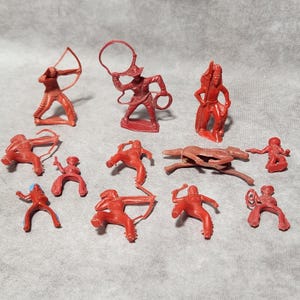May include: A collection of small, red plastic toy figures depicting cowboys, Native Americans, and a horse. The figures are in various action poses, including shooting bows and throwing lassos. The toys are likely from a vintage playset.