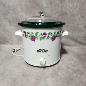 May include: White Rival Crock-Pot stoneware slow cooker with a clear glass lid and green trim. The slow cooker has a floral design of purple grapes and green leaves. The control knob has settings for Off, Low, and High.