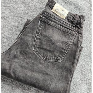 May include: Faded black denim jeans with a classic five-pocket design. The jeans have a back pocket with a stitched design and a leather patch. The jeans are folded, displaying the back pocket and waistband.