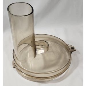 Peut inclure: Couvercle de robot culinaire en plastique transparent teinté, avec un tube d'alimentation cylindrique. Le couvercle est rond avec un bord légèrement surélevé et une petite languette sur le côté. Le plastique est translucide.