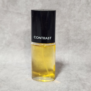 May include: A perfume bottle with a black cap and clear glass body. The bottle contains a yellow liquid. The word "CONTRAST" is printed in white on the black cap. The bottle is on a light grey background.