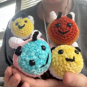 May include: Four handmade crochet bee toys in various colors: yellow, orange, blue, and yellow. Each bee has black eyes, a smiling mouth, and white wings. The bees are soft and plush, perfect for children or as decorative items.