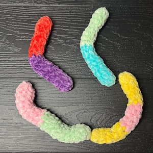 May include: Four colorful, plush, worm-shaped toys in red, purple, green, blue, pink, and yellow. The soft, textured toys are arranged on a dark wooden surface. These handmade toys are suitable for children.