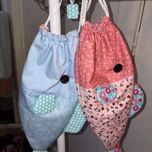 May include: Two handmade drawstring bags shaped like fish, hanging from a white bar. One bag is light blue with white polka dot accents, the other is pink with a floral pattern. Both have black button eyes and white rope drawstrings.
