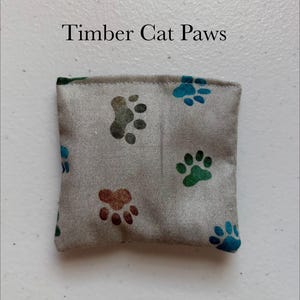 May include: A small, square, gray fabric item with colorful paw prints in shades of brown, green, and blue. The text "Timber Cat Paws" is printed in black on the top. The item appears to be a cat toy.