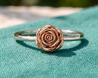 May include: A silver ring with a rose-shaped centerpiece. The rose is a warm, metallic color, and the band is smooth and shiny. The ring is set against a teal background, with sunlight creating shadows.