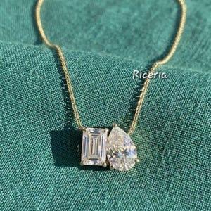 May include: A gold necklace featuring two diamond pendants: an emerald cut and a pear-shaped stone. The necklace is displayed on a green fabric surface. The word "Riceria" is visible.