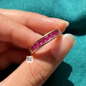 May include: A gold ring featuring a row of square-cut, vibrant pink gemstones. The ring is held against a teal background. The ring's design is simple, with the gemstones set closely together.