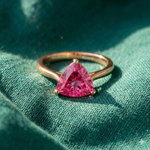 May include: A gold ring with a large, triangular, pink gemstone. The ring is set on a green fabric background. The word "Riceria" is visible on the ring.