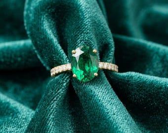 May include: A gold ring with a large, oval-cut emerald gemstone. The band is adorned with small, sparkling diamonds. The ring is set against a backdrop of deep green velvet.