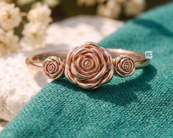 May include: A rose gold ring featuring three rose-shaped designs. The central rose is larger, with two smaller roses on either side. The ring is set against a teal fabric background, with a blurred floral backdrop.