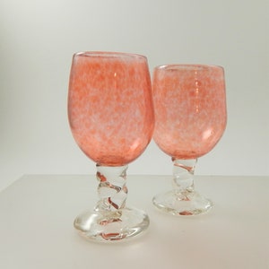 Peut inclure: Deux verres à vin à pied avec un bol tacheté de couleur corail et une tige torsadée transparente. La base des verres est transparente et circulaire. Les verres sont posés sur un fond blanc.