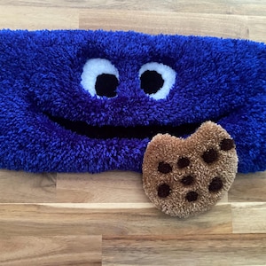 May include: A plush, blue, rectangular rug with a smiling face, white eyes, and a black mouth. A brown, plush cookie with chocolate chips is next to the rug. The rug and cookie are on a wooden surface.