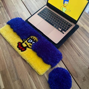 May include: A laptop with a screen displaying Marge Simpson, next to a yellow and blue rug with a cartoon character design, and a blue coaster. The items are on a wooden surface.