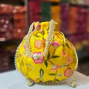 Può includere: Pochette gialla con coulisse con ricami floreali rosa e arancioni, accentuati da foglie verdi. La pochette è adornata da un cinturino di perle e una grande perla al centro. Nappe dorate pendono dal basso.