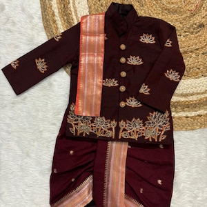 Puede incluir: Conjunto tradicional burdeos para niño, con una chaqueta con bordados de loto dorados y botones dorados, combinada con un pantalón a juego. Una bufanda decorativa con detalles en naranja y dorado completa el conjunto. Presentado sobre una superficie blanca.