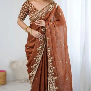Könnte beinhalten: Ein brauner Sari mit aufwendiger Goldstickerei. Der Sari hat eine passende Bluse mit kurzen Ärmeln und einem V-Ausschnitt. Die Goldstickerei befindet sich an den Rändern und auf der Bluse und sorgt für einen luxuriösen Look. Der Stoff scheint ein leichtes, fließendes Material zu sein.