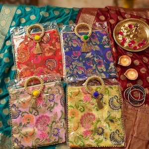 Könnte beinhalten: Vier farbenfrohe Stofftaschen mit floralen Mustern, goldenen Griffen und Quastenakzenten. Die Taschen sind in Rot, Blau, Rosa und Gelb gehalten und werden auf gemustertem Stoff präsentiert. Eine dekorative Platte mit Blumen und Kerzen ist ebenfalls zu sehen.
