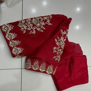 May include: A vibrant red blouse with intricate gold embroidery. The embroidery features floral and leaf patterns along the sleeves and neckline. The blouse is made of a smooth, possibly satin-like fabric.