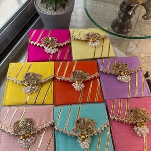 May include: A selection of vibrant pouches in various colours, including pink, yellow, orange, and teal. Each pouch is adorned with gold detailing, vertical stripes, and an elephant-shaped ornament with hanging pearls. The pouches are displayed on a white surface.