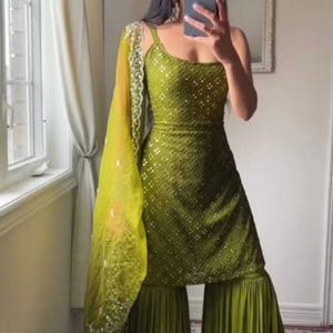 Puede incluir: Traje Sharara verde oliva con un top ajustado y adornado y pantalones de pernera ancha. El conjunto incluye una dupatta a juego con detalles bordados. Una versión contemporánea del atuendo tradicional indio, adecuado para ocasiones festivas.