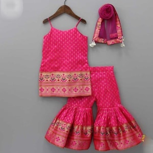 May include: A vibrant pink outfit for a child, featuring a camisole top, flared pants, and a matching scarf. The set is adorned with a decorative gold and brown border. The outfit is hanging on a wooden hanger against a gray background.