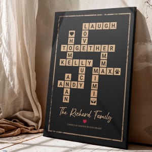 May include: Black framed family sign with a crossword design. The crossword includes the words "LAUGH, TOGETHER, KELLY, ANDY, MAX" and family names. The sign also includes the text "The Richard Family" and "FAMILY IS ALWAYS IN OUR HEART".