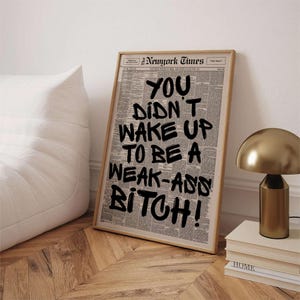 May include: Framed print featuring a newspaper background with the text "YOU DIDN'T WAKE UP TO BE A WEAK-ASS BITCH!" in bold black lettering. The print is in a light wood frame, leaning against a white wall.
