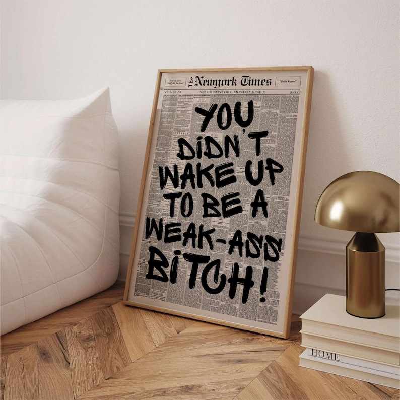 May include: Framed print featuring a newspaper background with the text "YOU DIDN'T WAKE UP TO BE A WEAK-ASS BITCH!" in bold black lettering. The print is in a light wood frame, leaning against a white wall.