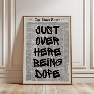 Puede incluir: Una l&aacute;mina de arte mural enmarcada con un fondo de peri&oacute;dico antiguo titulado The Black Times. Superpuesto en grandes letras negras en negrita con trazos de pincel aparece el texto JUST OVER HERE BEING DOPE. Marco de madera clara.