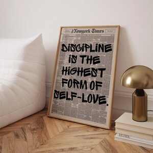 May include: A framed print featuring a newspaper background with the text "DISCIPLINE IS THE HIGHEST FORM OF SELF-LOVE" in bold black letters. The print is in a light wooden frame, leaning against a white wall.