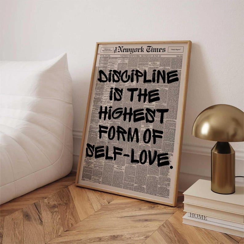 May include: A framed print featuring a newspaper background with the text "DISCIPLINE IS THE HIGHEST FORM OF SELF-LOVE" in bold black letters. The print is in a light wooden frame, leaning against a white wall.