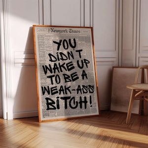 May include: A framed print featuring a newspaper background with bold black text that reads "YOU DIDN'T WAKE UP TO BE A WEAK-ASS BITCH!". The frame is a warm brown color, and the print is leaning against a white wall.