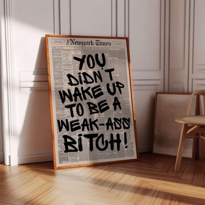 May include: A framed print featuring a newspaper background with bold black text that reads "YOU DIDN'T WAKE UP TO BE A WEAK-ASS BITCH!". The frame is a warm brown color, and the print is leaning against a white wall.