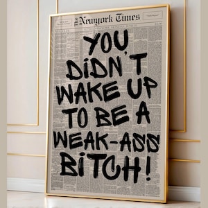 May include: A framed print with a newspaper background, the headline reads "The Newyork Times." The bold black text states "YOU DIDN'T WAKE UP TO BE A WEAK-ASS BITCH!" The print is enclosed in a gold frame.