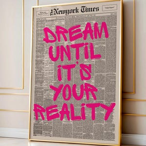 May include: A framed print featuring a vintage New York Times newspaper page. Bold, hot pink text reads "DREAM UNTIL IT'S YOUR REALITY." The newspaper is dated June 21 and has a gold frame.