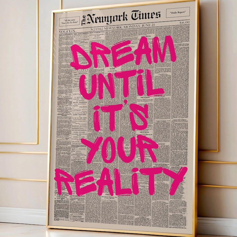 May include: A framed print featuring a vintage New York Times newspaper page. Bold, hot pink text reads "DREAM UNTIL IT'S YOUR REALITY." The newspaper is dated June 21 and has a gold frame.