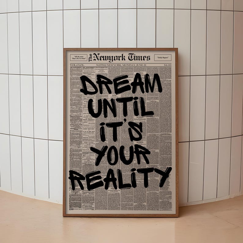 May include: A framed print of a vintage New York Times newspaper page. Bold black text overlays the newsprint, reading "DREAM UNTIL IT'S YOUR REALITY." The frame is a warm brown, and the background is a white tiled wall.