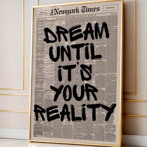 May include: A framed print featuring a vintage newspaper background with the words "DREAM UNTIL IT'S YOUR REALITY" in bold black lettering. The print is in a gold frame, adding a touch of elegance to the motivational message.