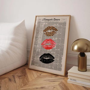 May include: Framed print featuring three sets of lips in leopard print, red, and black, set against a newspaper background. The top of the print reads "New York Times". The artwork is displayed in a light-colored wooden frame.