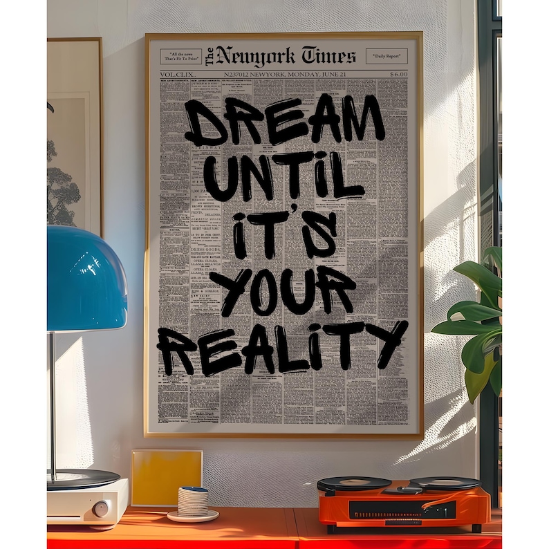 May include: A framed print of a New York Times newspaper page with the words "DREAM UNTIL IT'S YOUR REALITY" in bold black letters. The print is in a gold frame and is displayed on a wall. A blue lamp and orange record player are on a red table.