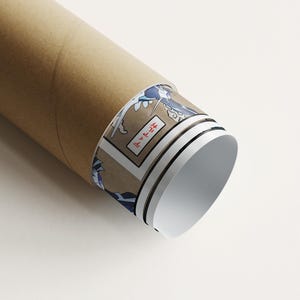 May include: A rolled-up poster in a brown cardboard tube. The poster's edge reveals a Japanese-style illustration with blue waves and text in a rectangular frame. The tube is on a white surface.