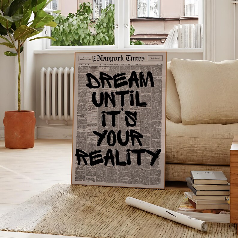 May include: A framed print of a New York Times newspaper page with the words "DREAM UNTIL IT'S YOUR REALITY" in bold black letters. The print is displayed on a rug in front of a window and a beige sofa.