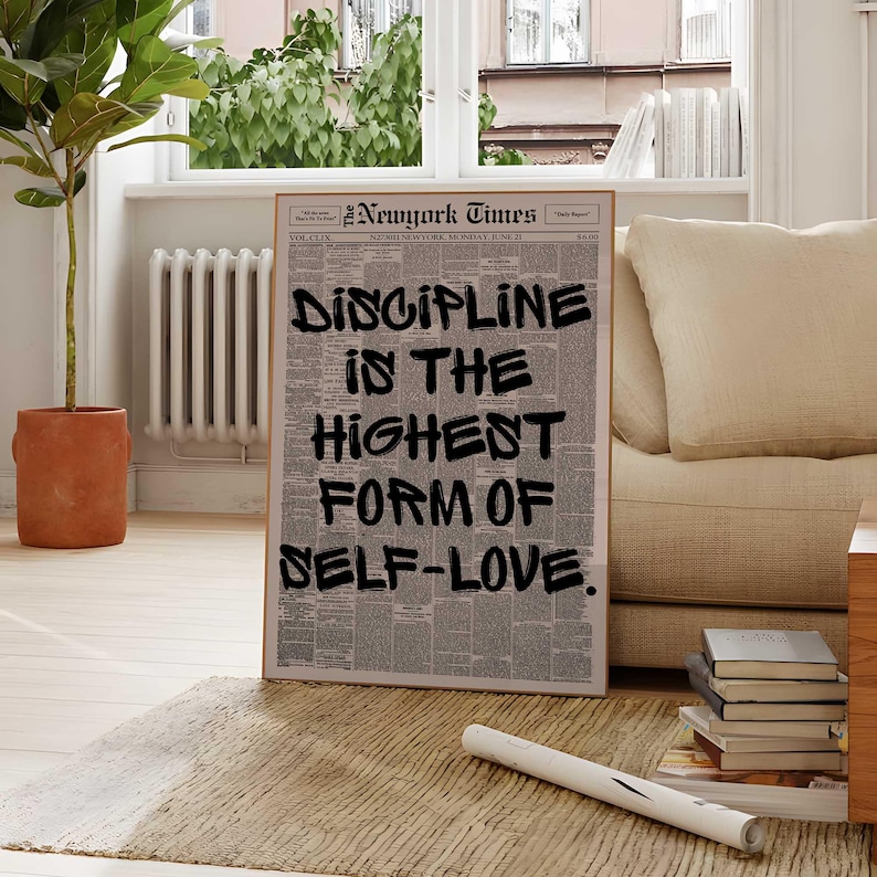 May include: A framed print of a New York Times newspaper page with the words "DISCIPLINE IN THE HIGHEST FORM OF SELF-LOVE" in bold black letters. The print is in a room with a beige sofa, a potted plant, and a window.