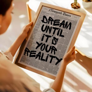 May include: A framed print featuring a newspaper page with the headline "The New York Times." Bold black text reads "DREAM UNTIL IT'S YOUR REALITY." The frame is made of light-colored wood, and the print is held by a person.