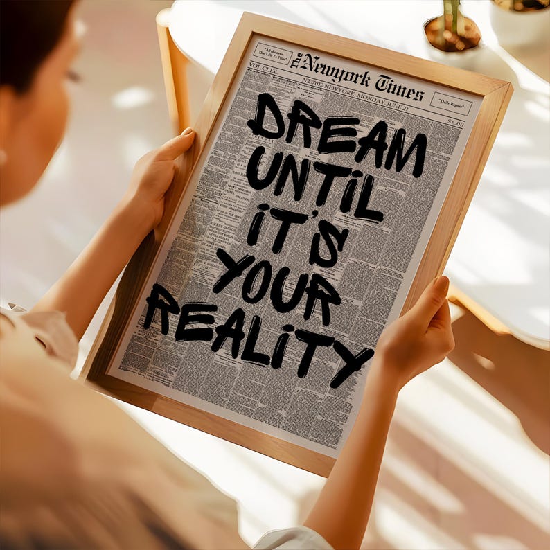 May include: A framed print featuring a newspaper page with the headline "The New York Times." Bold black text reads "DREAM UNTIL IT'S YOUR REALITY." The frame is made of light-colored wood, and the print is held by a person.