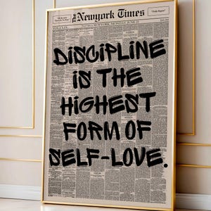 May include: A framed print featuring a vintage newspaper background with the headline "New York Times." Bold black text reads "DISCIPLINE IS THE HIGHEST FORM OF SELF-LOVE." The print is encased in a gold frame.