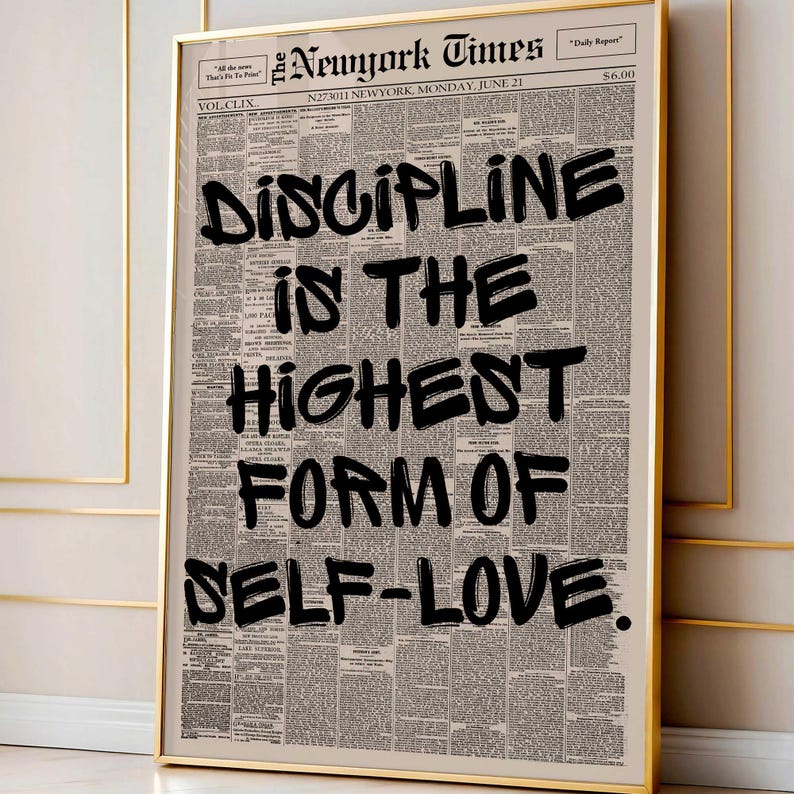 May include: A framed print featuring a vintage newspaper background with the headline "New York Times." Bold black text reads "DISCIPLINE IS THE HIGHEST FORM OF SELF-LOVE." The print is encased in a gold frame.