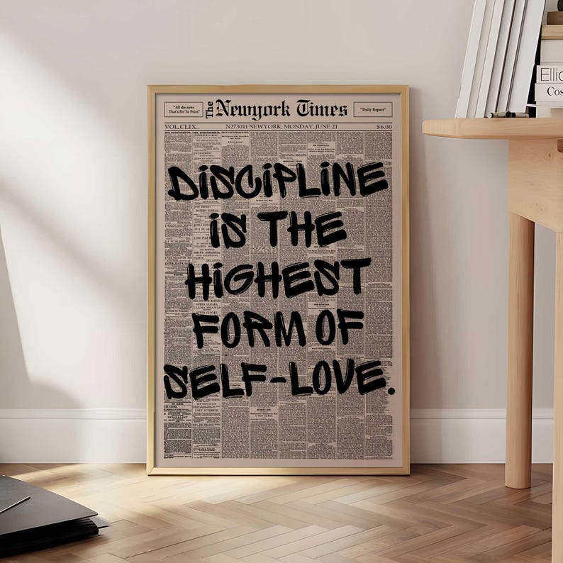 May include: A framed print featuring a vintage newspaper background. Bold black text reads "DISCIPLINE IS THE HIGHEST FORM OF SELF-LOVE." The print is in a light wood frame, and the newspaper is dated June 21.