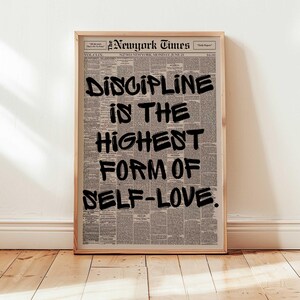May include: A framed print featuring a vintage New York Times newspaper page. Bold black text reads "DISCIPLINE IS THE HIGHEST FORM OF SELF-LOVE." The print is in a light wooden frame, displayed against a white wall and wooden floor.